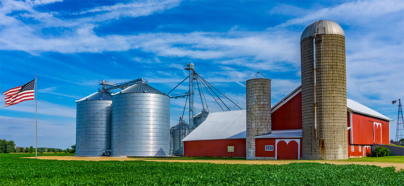 Barn with silos and storage bins on a summer day with big blue sky and American Flag blowing in the wind on a flag pole.
