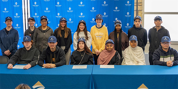 Signing Day students on the Faribault campus grouped together smiling and wearing SCC caps