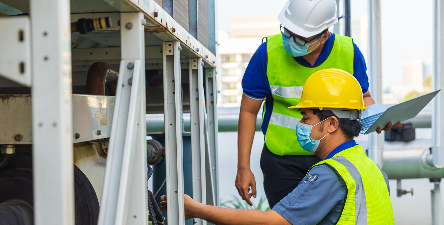 Two workers wearing hard hats and high-vis vests inspect industrial equipment