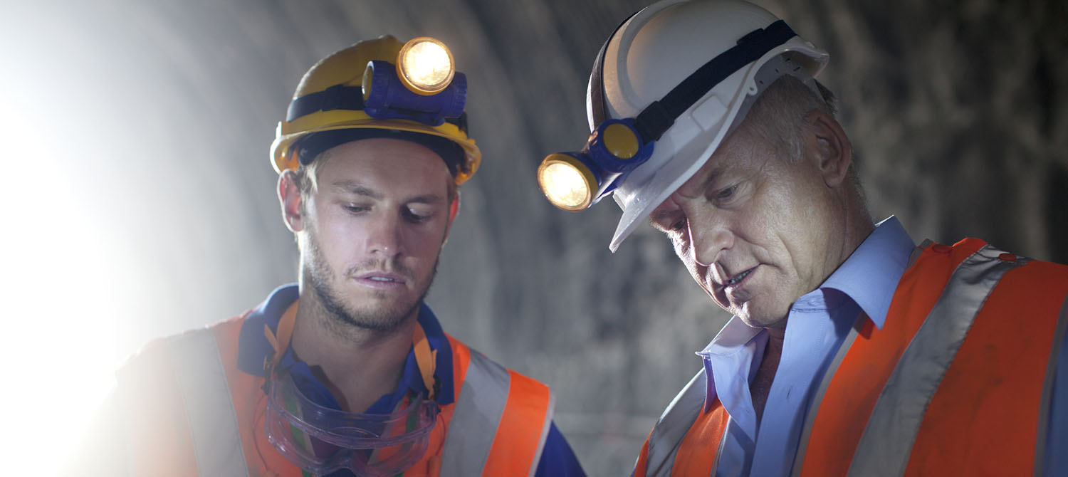 Two workers in a tunnel wearing hard hats with lights and high visibility vests talking to each other while looking off screen.