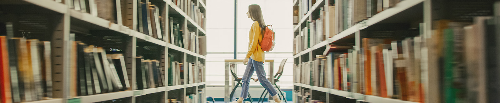 Student with a backpack walking by library shelves filled with books, heading toward a large window with natural light