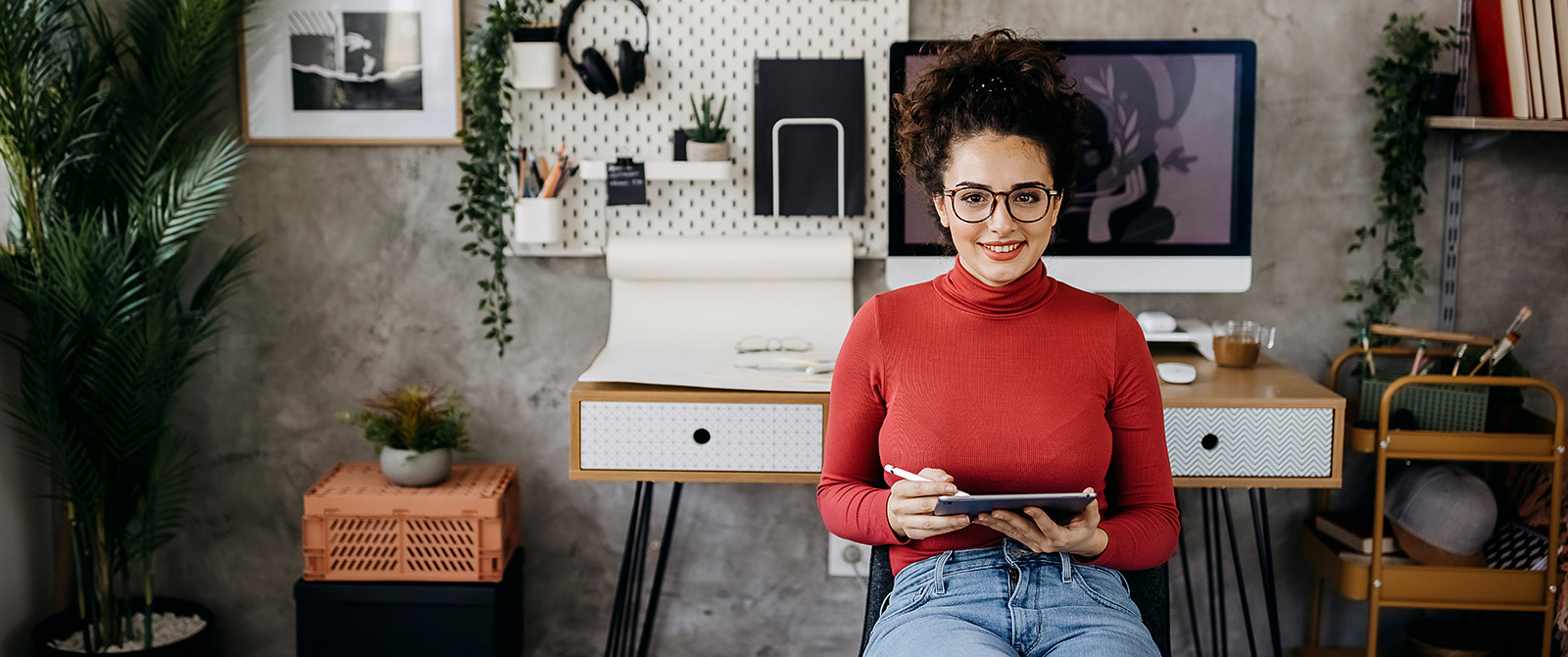 Young woman in her home office in front of computer and holding a tablet.