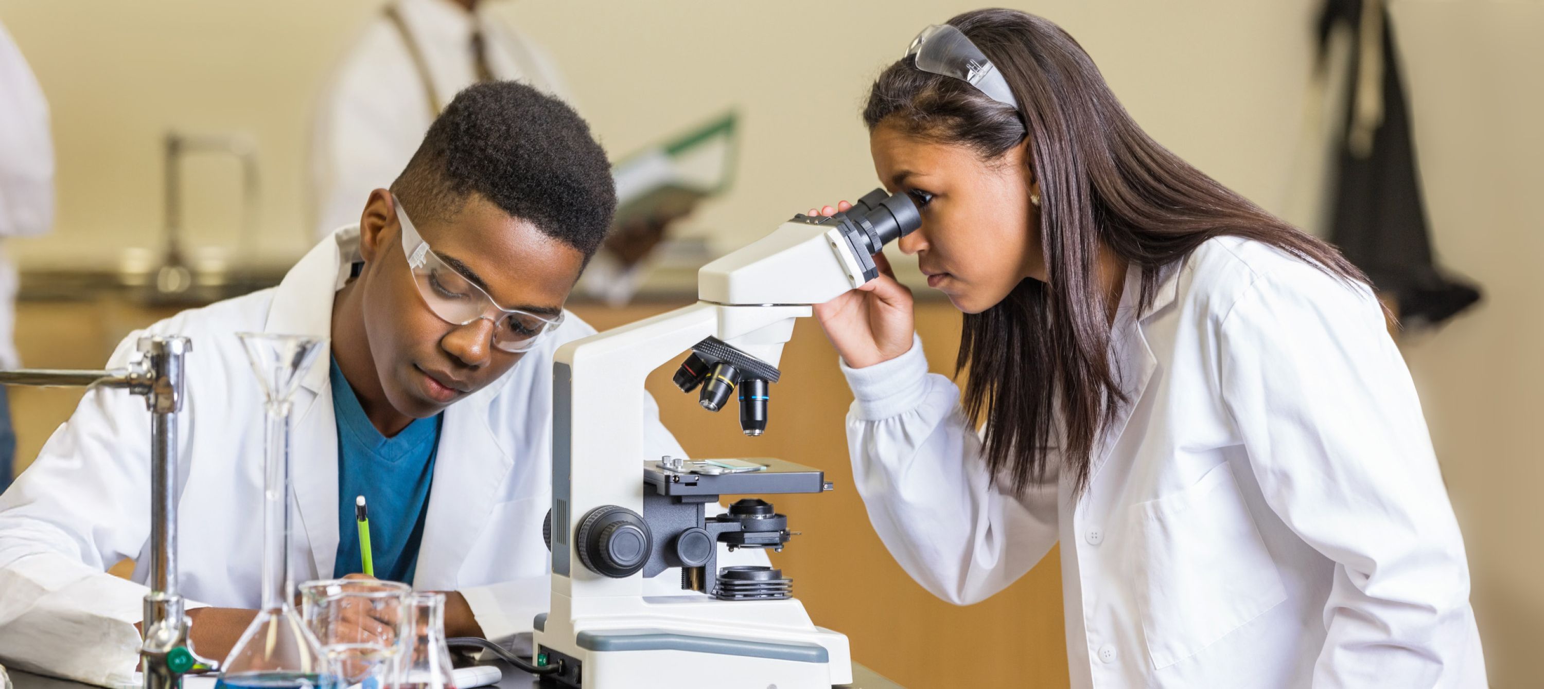 Two student working in a chemistry lab wearing white coats. One student is looking through a microscope while the other takes notes.