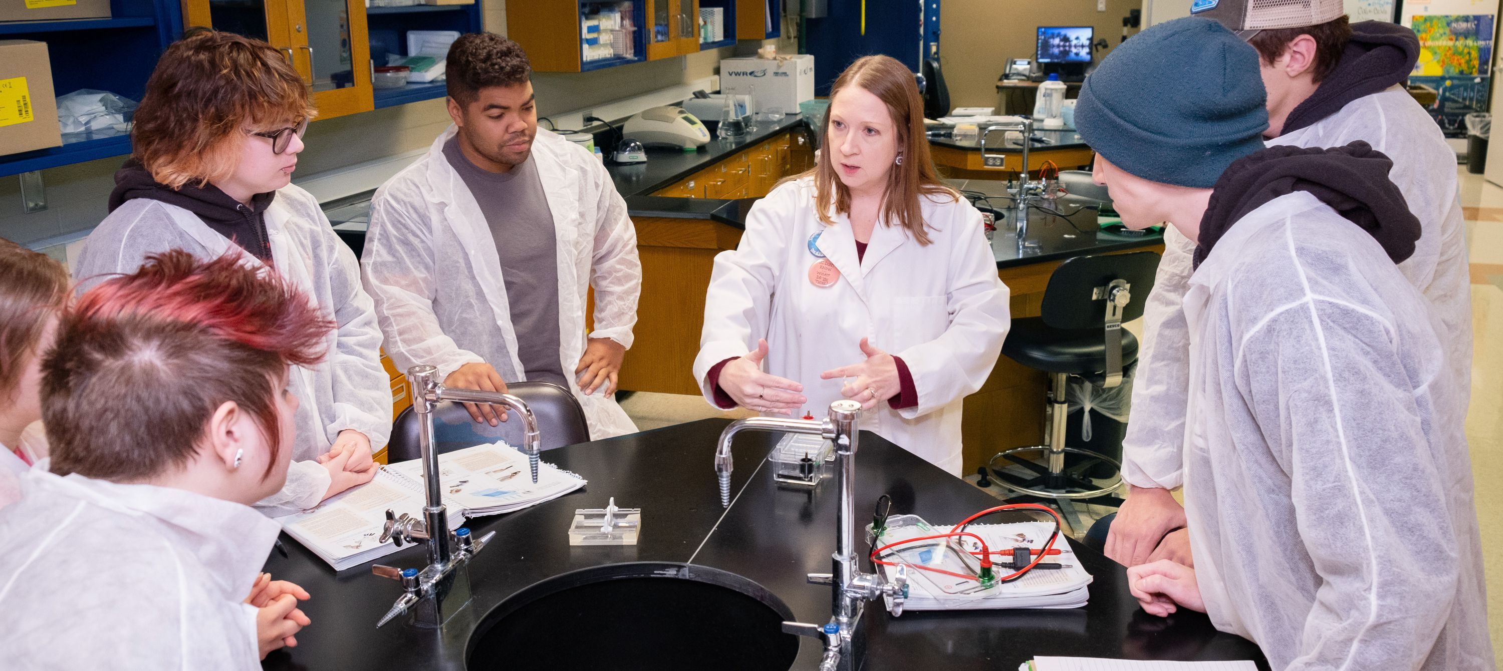 An instructor standing at a table in a biology lab interacting with a group of students.