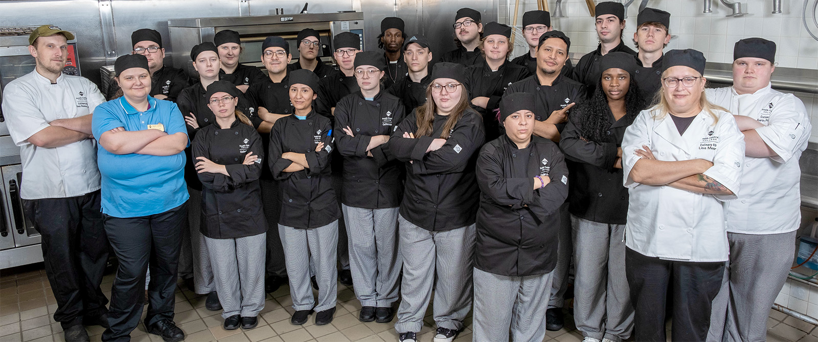 Large group of SCC culinary students and instructors in the culinary kitchen wearing uniforms and looking at the camera.