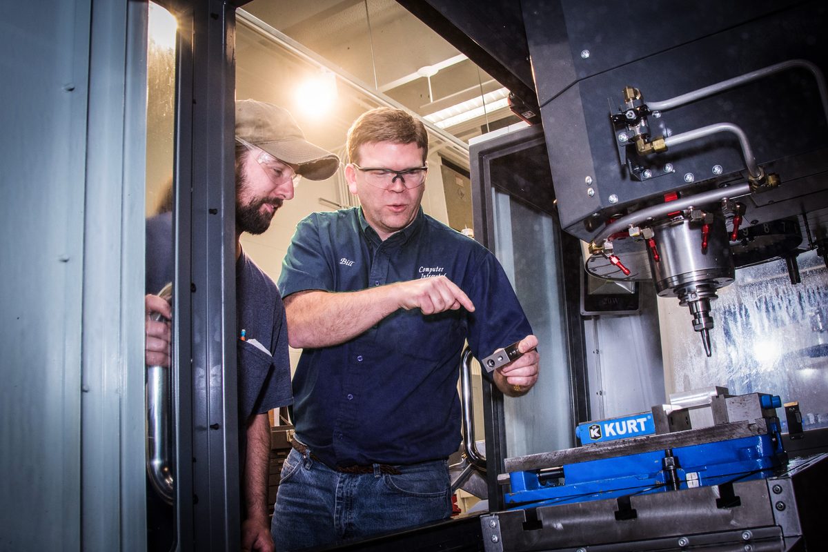 An experienced machinist points out a detail on a CNC mill while a student looks on.
