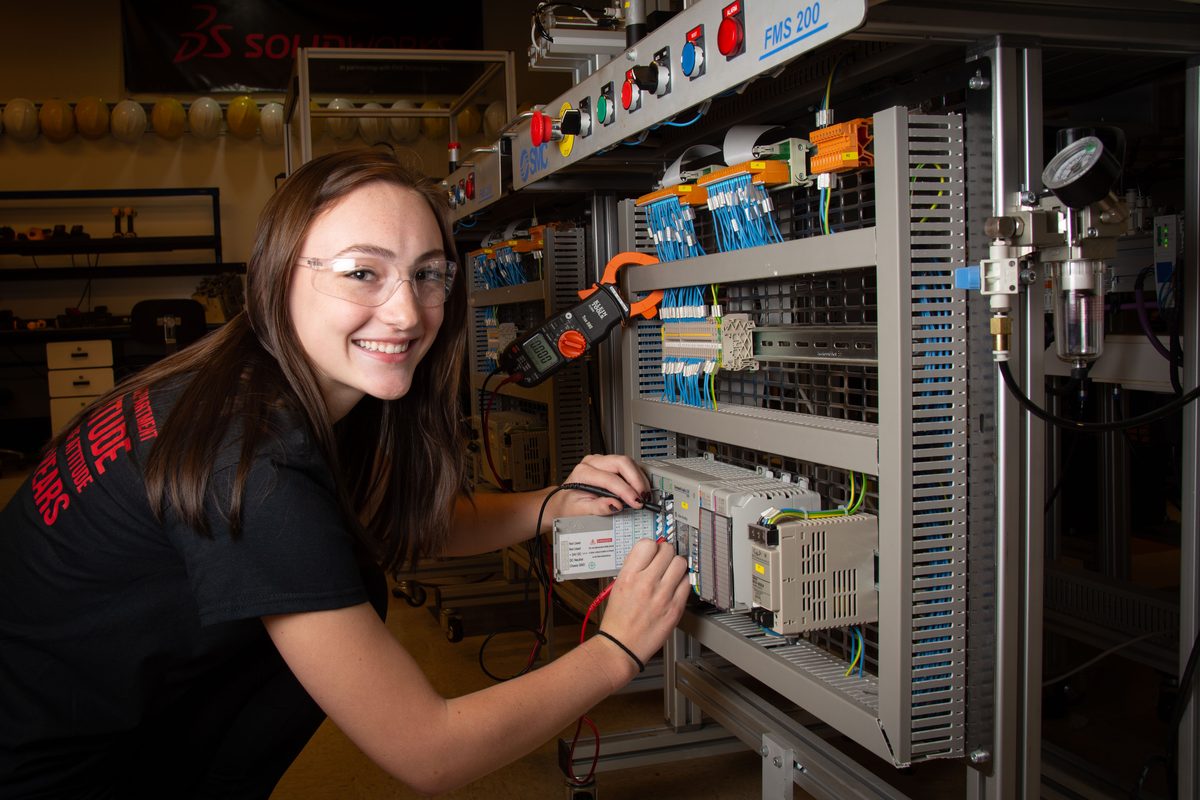 A smiling mechatronics student wearing safety glasses tests wiring on an industrial control panel.